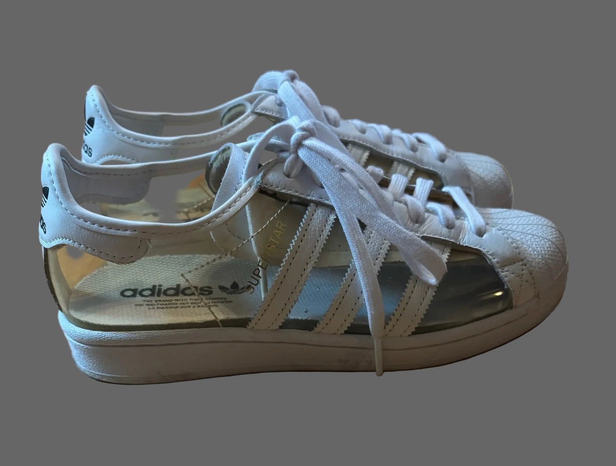 A pair of Adidas Superstar sneakers with clear plastic uppers and white suede overlays, resting on a wooden floor.