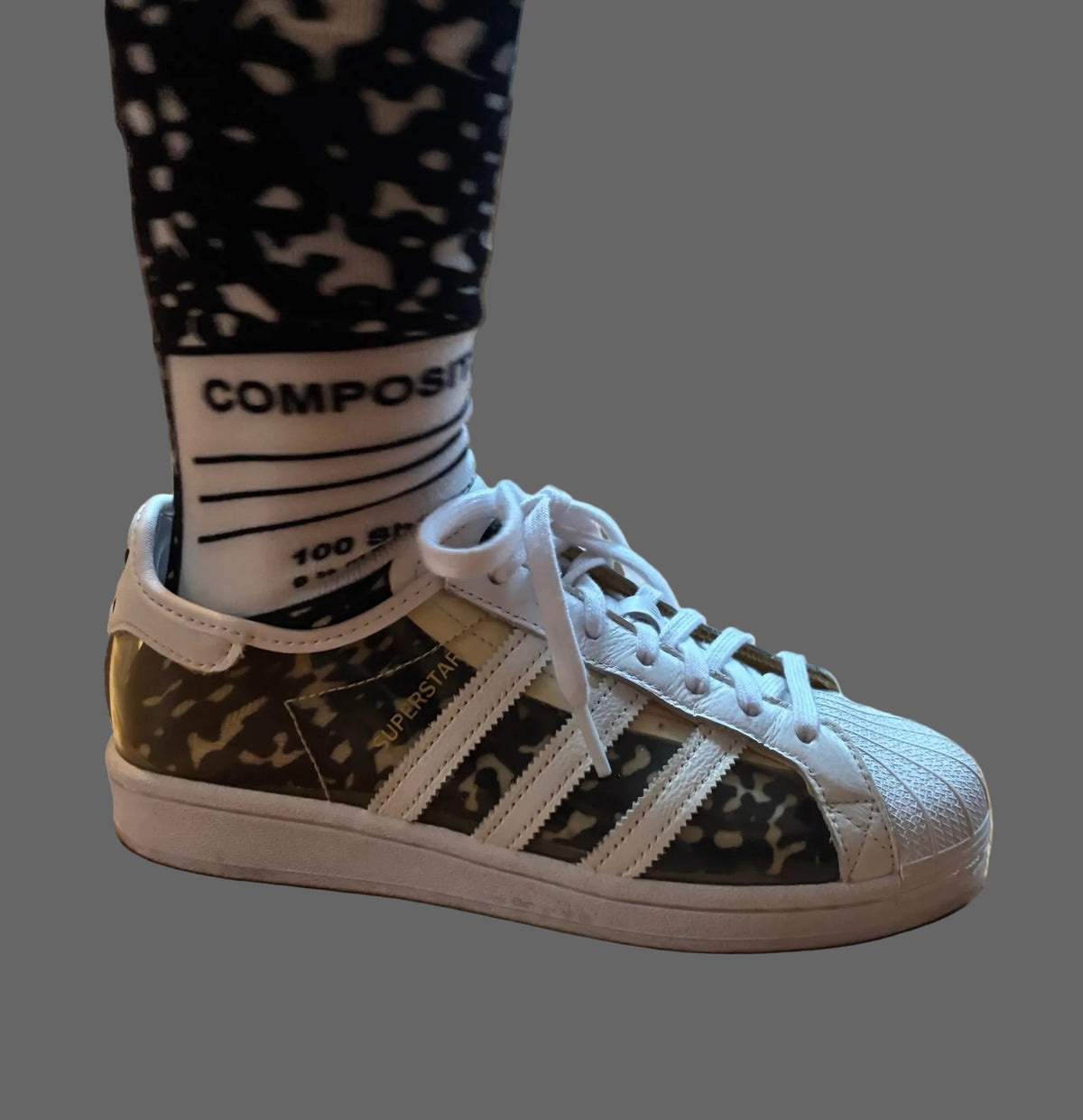 A person wearing a white Adidas Superstar sneaker with a green and black camouflage pattern on the side panels, paired with a black and white camo sock on a wooden floor.