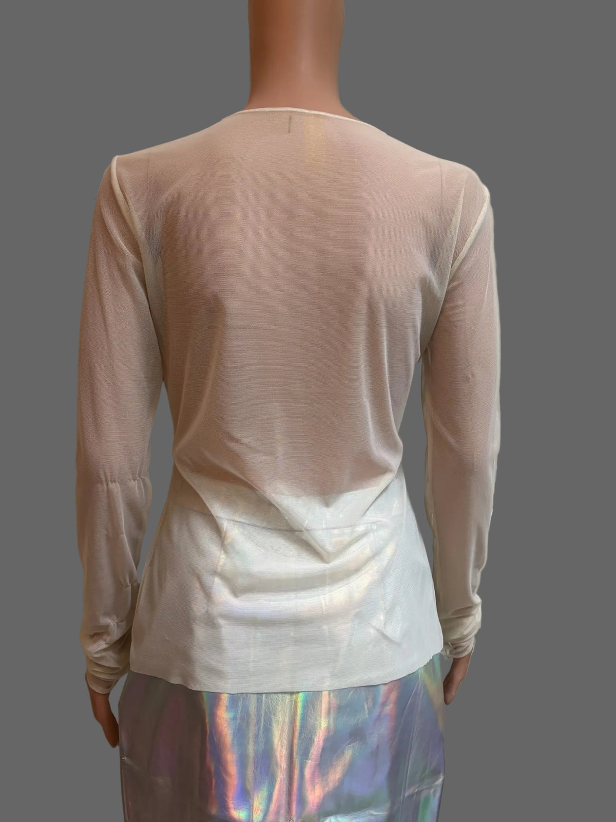 A long-sleeve sheer mesh top in a pale blush pink color, shown from the back on a mannequin. The top is layered over a shiny, iridescent holographic skirt or pants.