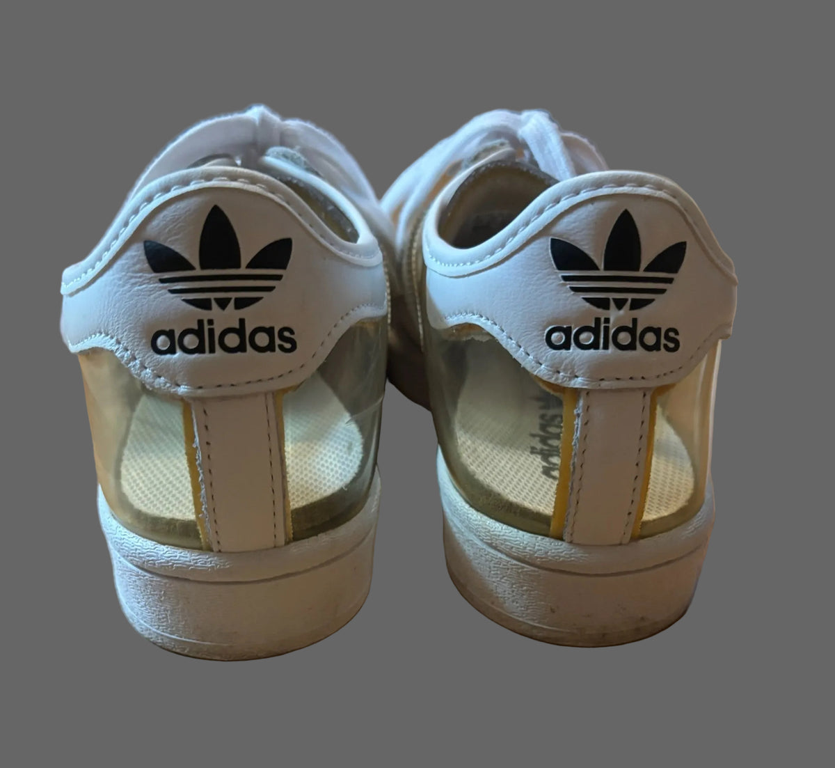 A pair of white Adidas Originals Superstar sneakers viewed from the back, resting on a wooden floor. The shoes feature the classic trefoil logo and brand name on the heel tab, with a translucent clear plastic side panel.