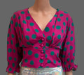A vintage-style blouse with a vibrant magenta pink base and teal green polka dots, styled on a mannequin with a shiny silver skirt against a white paneled door background.