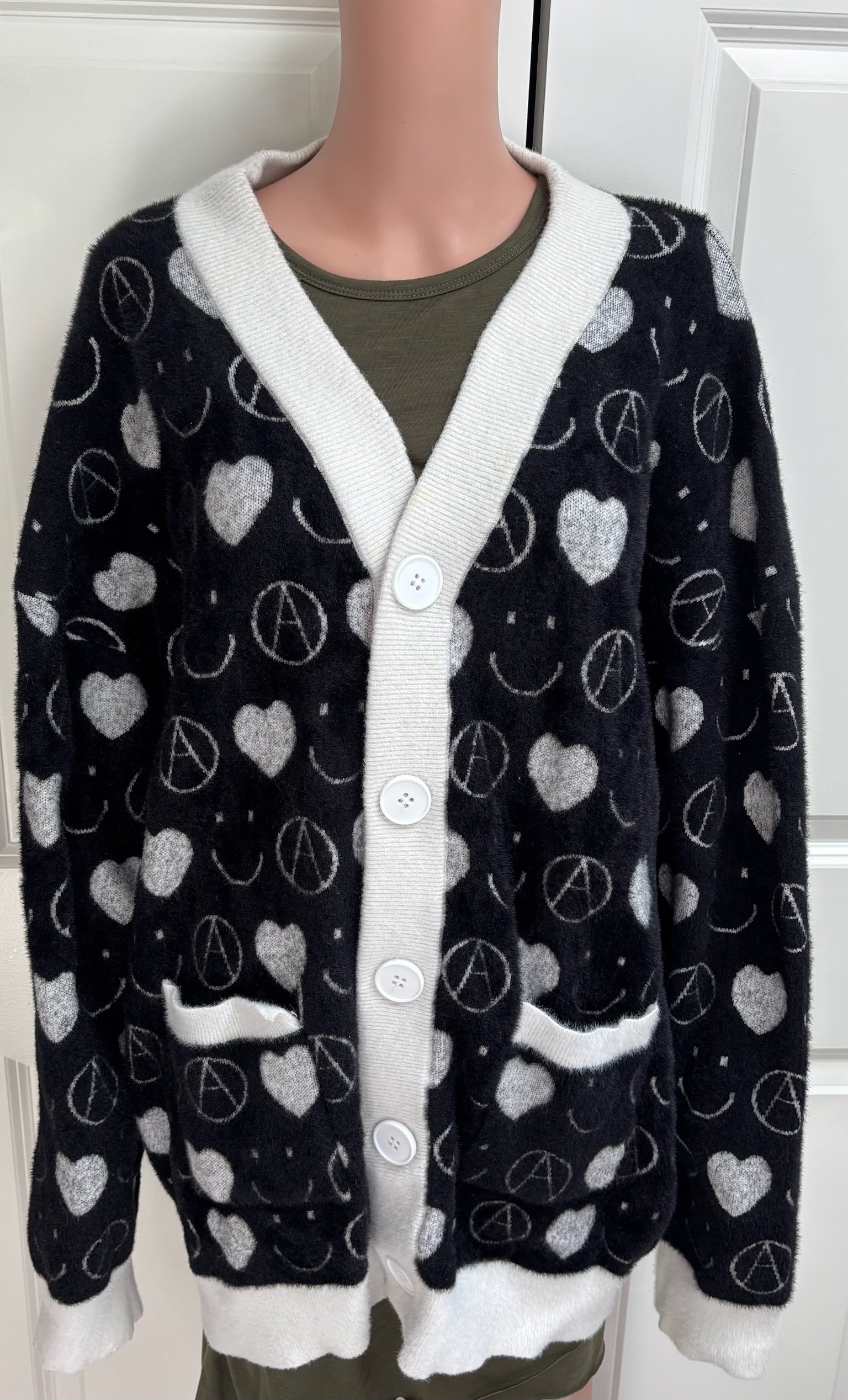 A black fuzzy cardigan sweater with white trim and a repeating pattern of white hearts, peace signs, and smiley faces, displayed on a mannequin torso.