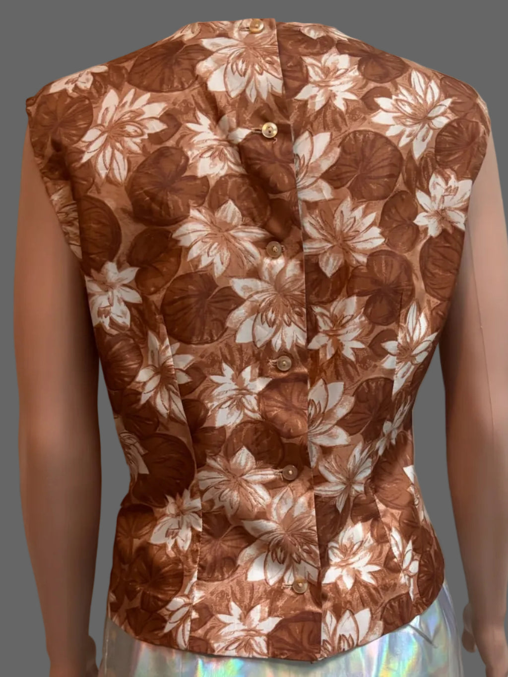 A sleeveless brown and beige floral vest displayed on a mannequin, paired with an iridescent holographic skirt, set against a white paneled door background.