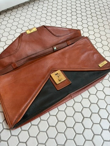 A brown leather envelope-style clutch or document bag with gold-tone hardware, shown partially open on a white hexagonal tile floor. A black interior lining is visible, along with a small leather brand patch and a gold tag.