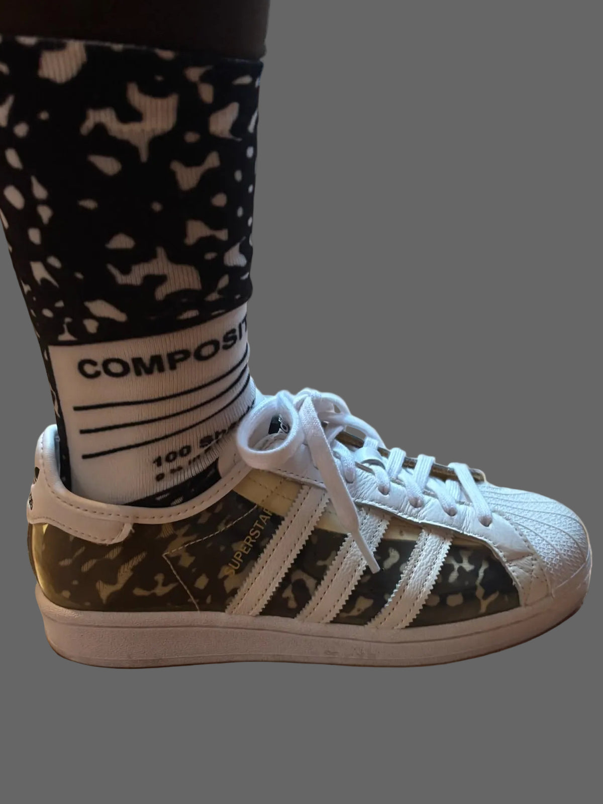 A person wearing a camouflage Adidas Superstar sneaker with white laces and a white sock featuring a black and white pattern, standing on a wooden floor.