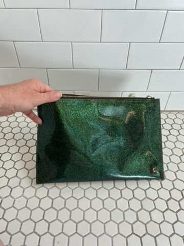 A hand holds a rectangular green glittery clutch bag with a gold zipper, set against a white tiled background.