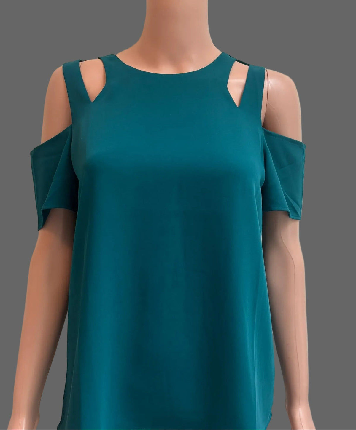 A teal cold-shoulder top displayed on a mannequin torso in front of a white paneled door. The top features cutout details at the shoulders and short draped sleeves.