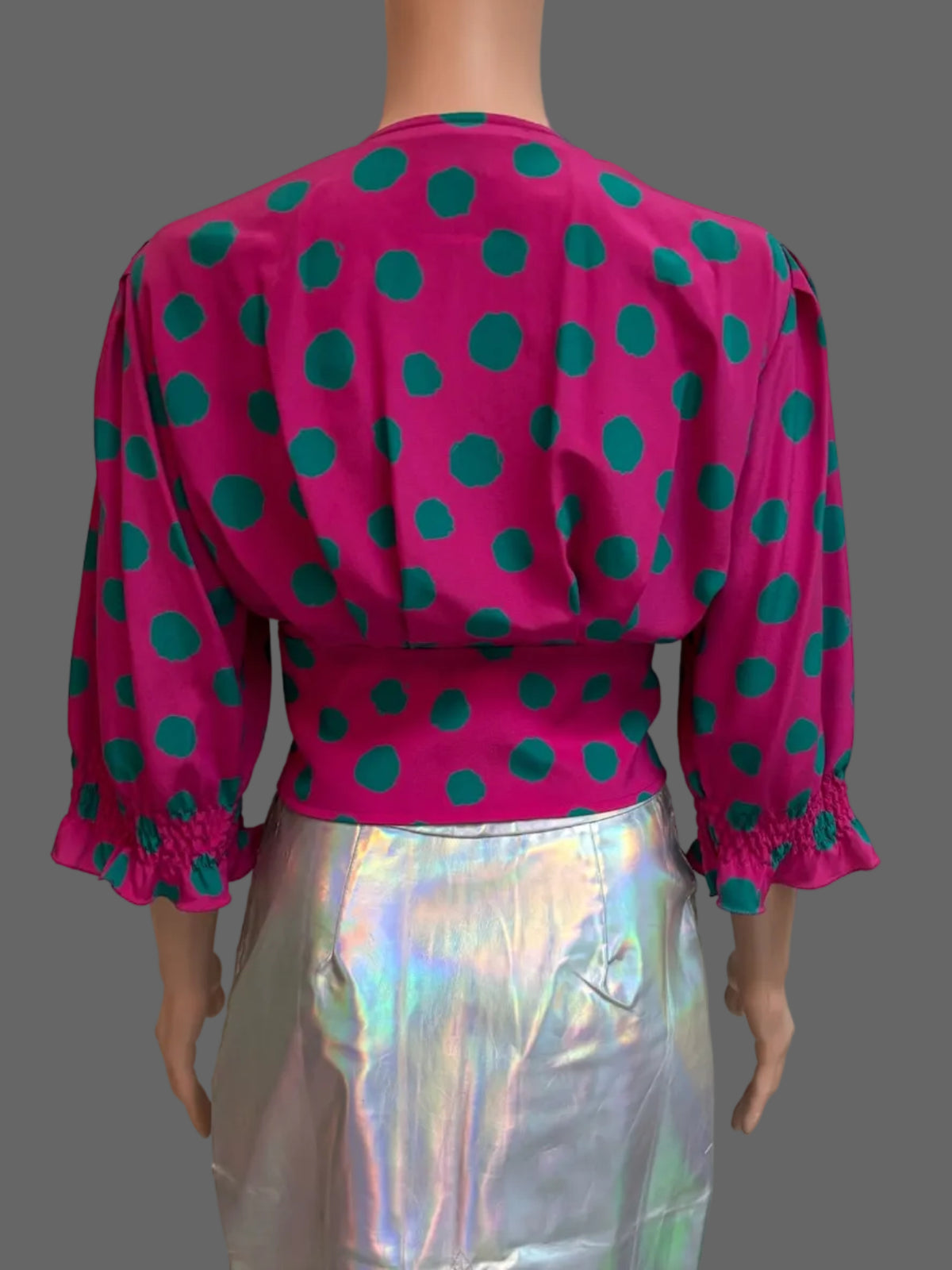 A mannequin wearing a bright pink blouse with teal green polka dots and a shiny holographic silver skirt, shown from the back in front of a white paneled door.