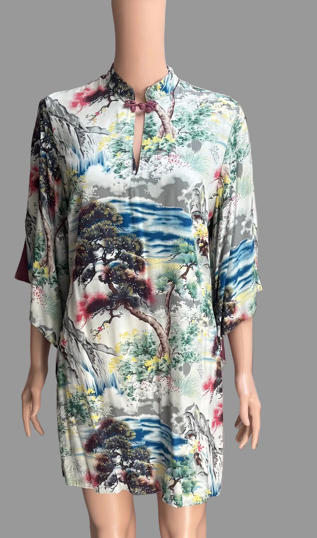 A short-sleeved, high-neck tunic dress with a colorful landscape print, displayed on a mannequin against a gray curtain backdrop.