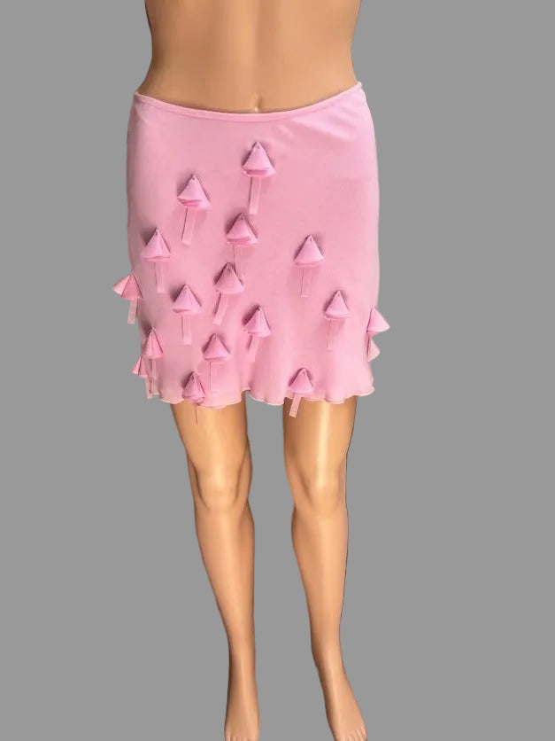 A pink mini skirt with a ruffled hem and small hanging pink bow accents, displayed on a mannequin in front of draped taupe curtains.