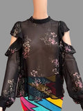 A sheer black long-sleeve blouse with floral print and cold-shoulder ruffle sleeves, displayed on a mannequin torso with a colorful skirt underneath, set against a brown curtain backdrop.