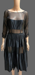 A black sheer mesh dress displayed on a mannequin in front of brown curtains. The dress features a square neckline, three-quarter sheer sleeves, a solid black bust panel, a sheer waist band, and a pleated skirt with sheer window panels.