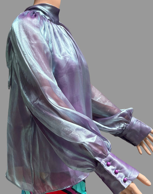 A sheer, iridescent lavender-purple blouse with a high mock neck and three-button cuffs, displayed on a mannequin against a brown curtain background.