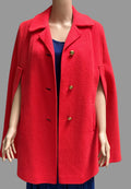 A bright red cape-style coat displayed on a mannequin, worn over a blue top and blue pleated skirt, against a gray curtain backdrop.